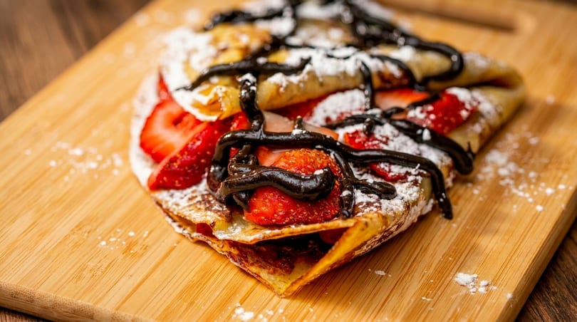 A strawberry and chocolate crepe, dusted with powdered sugar, on a wooden board.