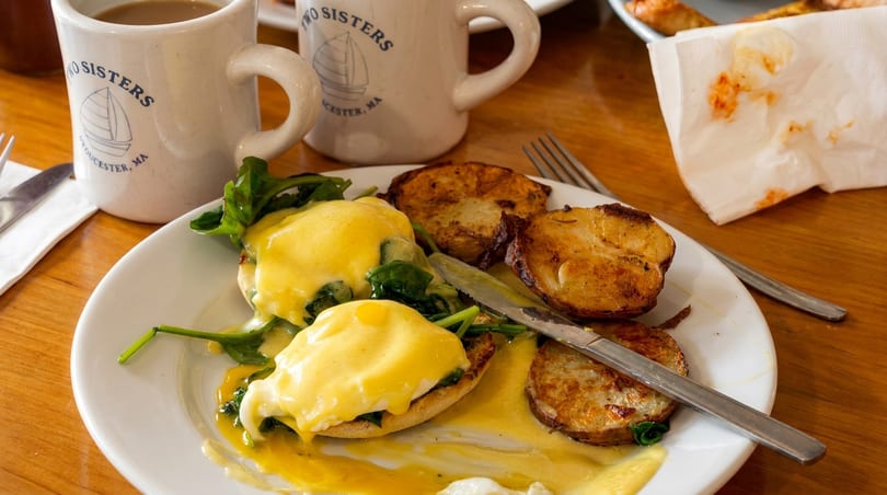 Eggs Benedict with spinach and roasted potatoes, with Two Sisters branded coffee mugs in the background.