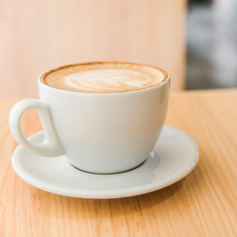 A white cup of latte with foam art sits on a matching saucer on a light wooden table.