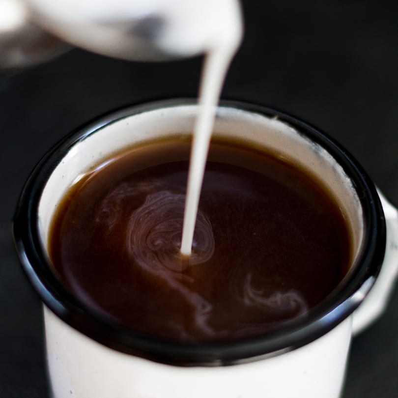 Milk pouring into a dark coffee in a white enamel mug, creating a swirl.